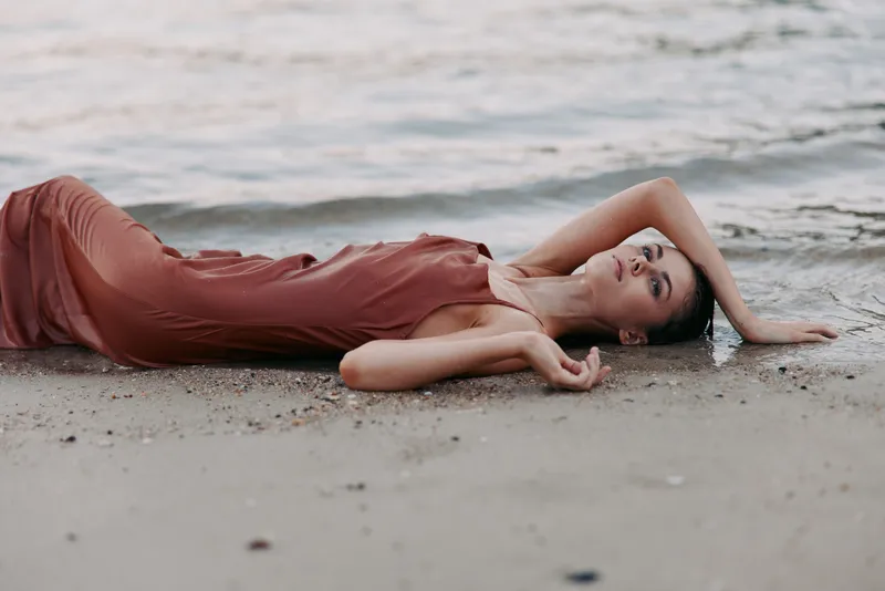 A woman lies on damp sand near the calm shoreline, draped in a flowing pink dress, with relaxed pose and soft lighting creating a serene beach scene.