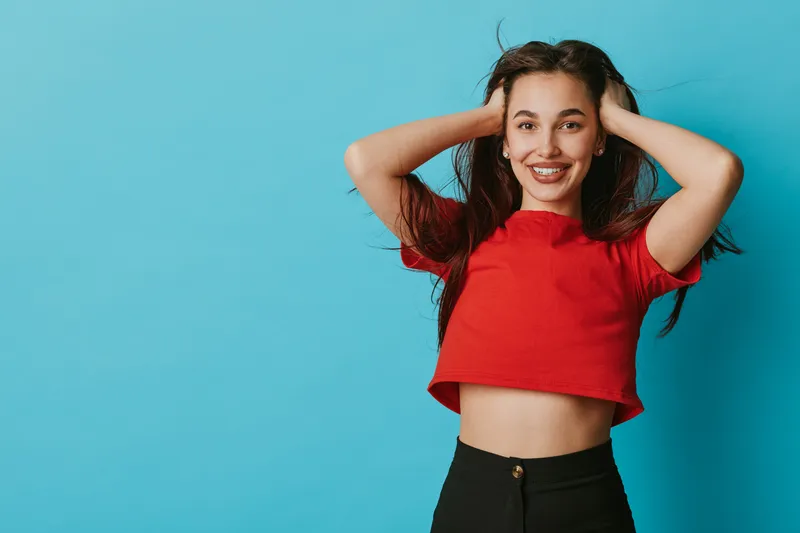 Young woman in red crop top smiling with hands in hair against blue background. Casual style, happiness and confidence in female portrait with natural expression and long dark hair.