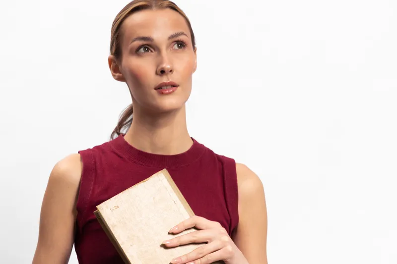 Thoughtful young woman in sleeveless red top holds closed book, looking away on white background. Concept of reading, education, and contemplation with soft natural lighting.