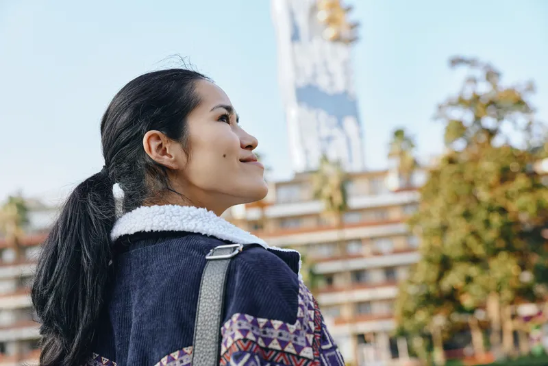 Woman with backpack outdoors, looking upward at the sky beside urban buildings, sunlit scene conveying curiosity, exploration and hope in a modern city environment