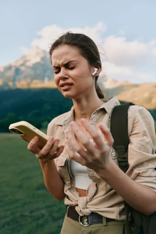 Young woman with backpack listens to music on wireless earbuds while holding smartphone outdoors in mountains, showing emotional reaction to sound, lifestyle concept
