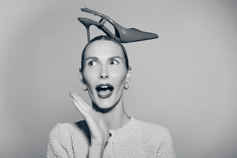 Surprised Caucasian woman with fair skin wearing earrings and sweater balances a high heel shoe on her head. Studio portrait with neutral background. Concept of fashion and humor.