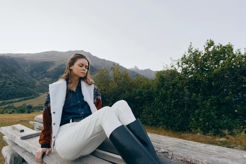 Woman portrait in mountains nature outdoors, fashion look with coat and boots, relaxed pose on wooden bench, scenic valley background, autumn travel and peaceful mood.