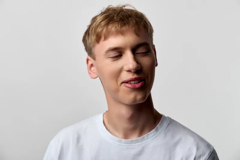 Man with a relaxed smile wearing a white t shirt, eyes closed, head tilted slightly, soft studio lighting, neutral gray backdrop, casual mood, a candid moment of ease and comfort.