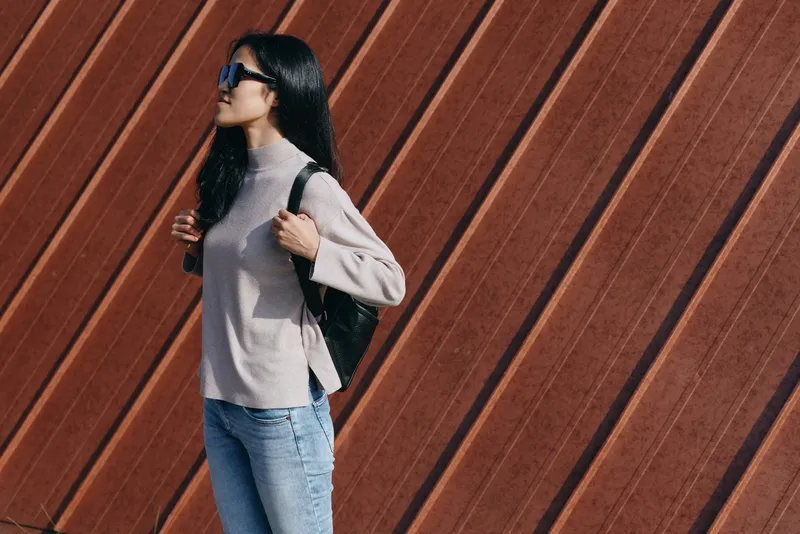 Woman wearing sunglasses and soft sweater with backpack walking outdoors along a diagonal wooden wall, showcasing casual fashion, travel vibe, urban style, daylight mood, relaxed attitude.