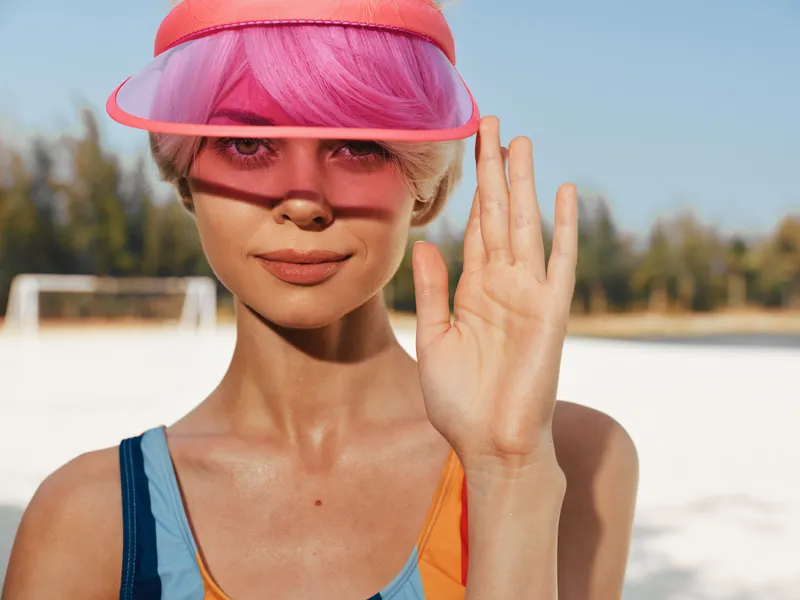 Young woman with pink hair under a translucent pink visor raises a hand in a sunny beach scene, wearing a bright swimsuit as warm light creates a vibrant, playful mood.