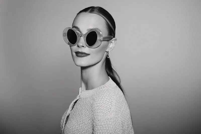 Elegant white woman with pale skin wearing round sunglasses and textured jacket poses confidently against plain background.