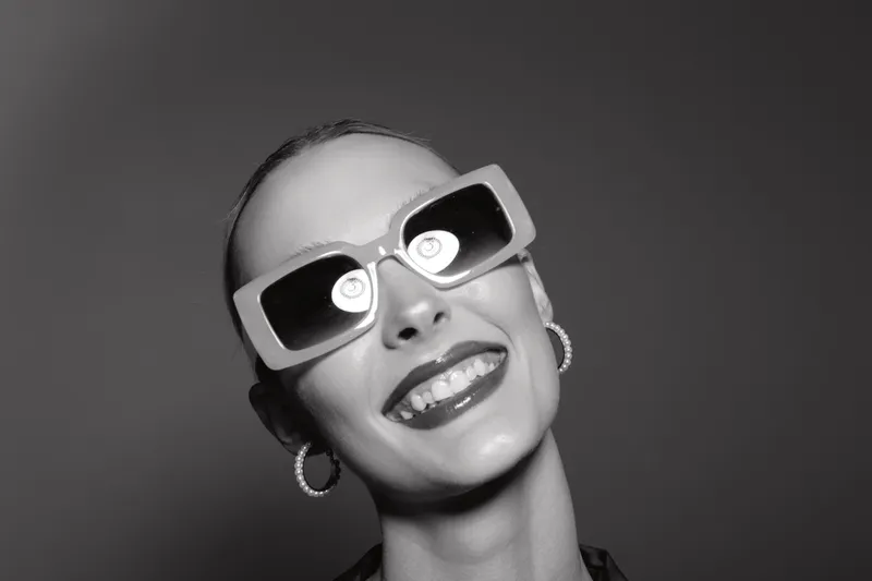 Smiling white woman with light skin wearing large sunglasses and hoop earrings poses on grey background. Concept for fashion, style, and confident beauty portrait.