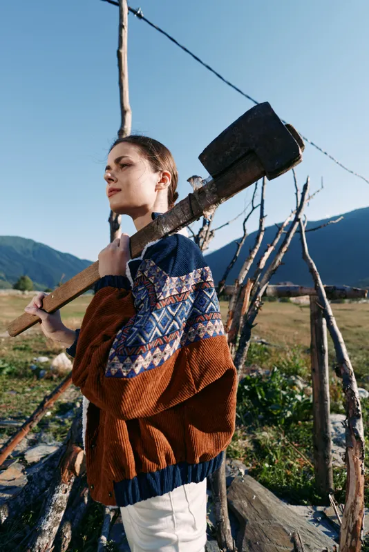 Woman carrying sledgehammer over shoulder on farm with mountains in background, wearing patterned sweater near wooden fence, rural outdoor portrait and hard work scene.