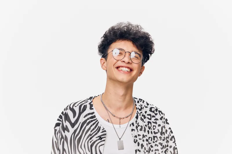 smiling young man wearing glasses with curly hair, dressed in patterned black and white shirt against a plain white background, joyful expression, casual style
