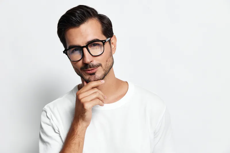 Thoughtful young man wearing glasses and a white t-shirt, posing with a hand on his chin, looking directly at the camera against a plain background. People lifestyle concept.