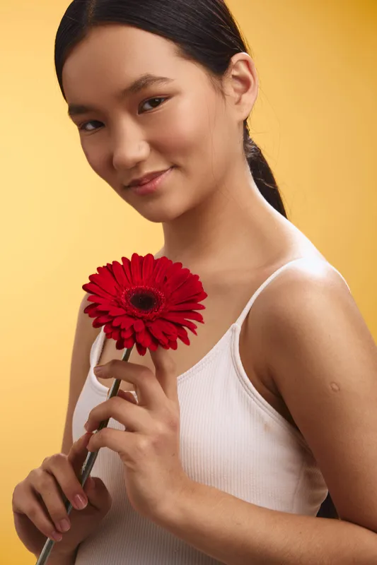 Asian woman holds a red flower against a warm yellow background, smiling softly. A graceful, natural portrait that conveys calm, elegance, and contemplative charm.