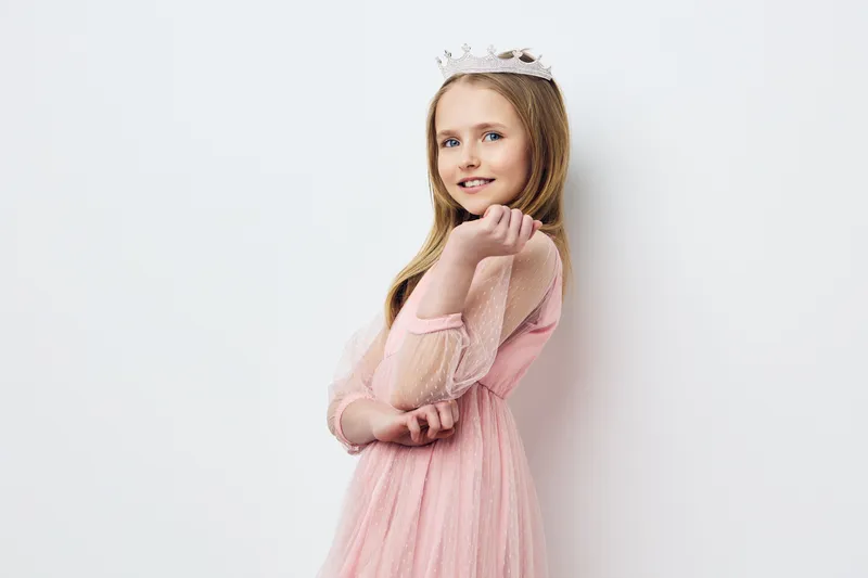 Young girl in a pink dress with a crown, smiling joyfully against a light background, reflecting a cheerful and playful mood