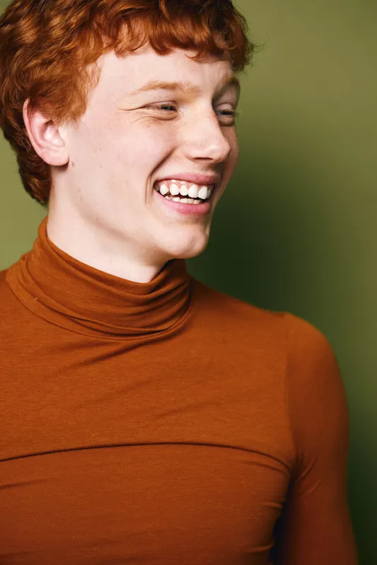 Young redhead man smiling happily in brown turtleneck sweater, looking aside with teeth showing, natural light and green background for casual portrait and positive emotion.