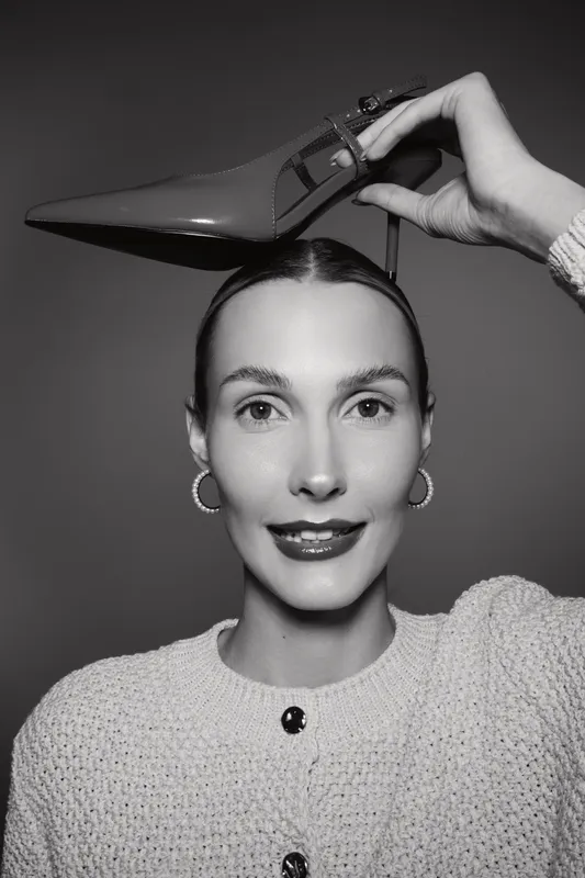 Young white woman with pale skin holds red high heel shoe above head. Studio portrait in black and white. Concept for fashion, style, and elegance.