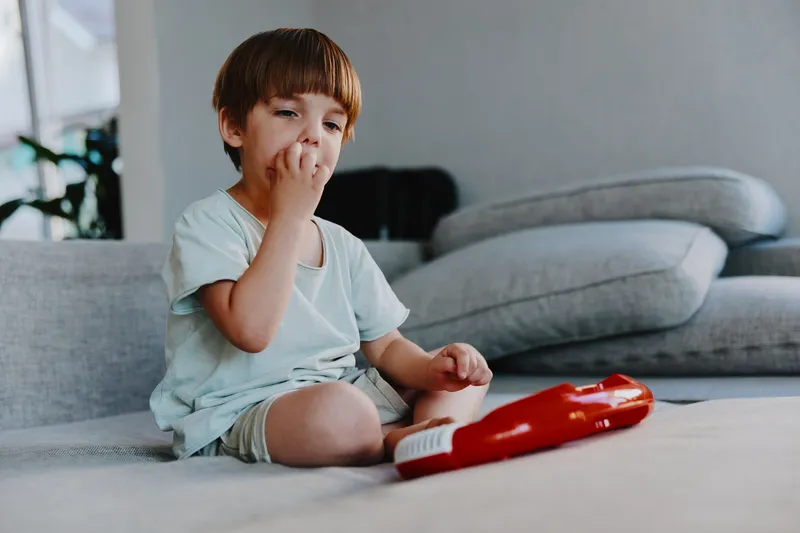 Thoughtful boy enjoying playtime, sitting on a cozy sofa with a red toy. The room is bright and inviting, adding to the cheerful atmosphere.