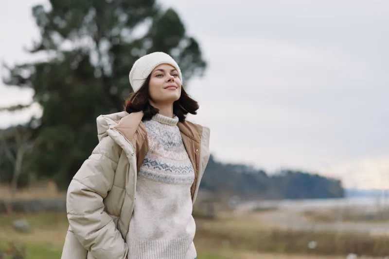 Woman outdoors in a winter coat and knitted sweater, wearing a beanie, smiles confidently in a breezy landscape. Natural light highlights fashion and relaxed lifestyle in nature.