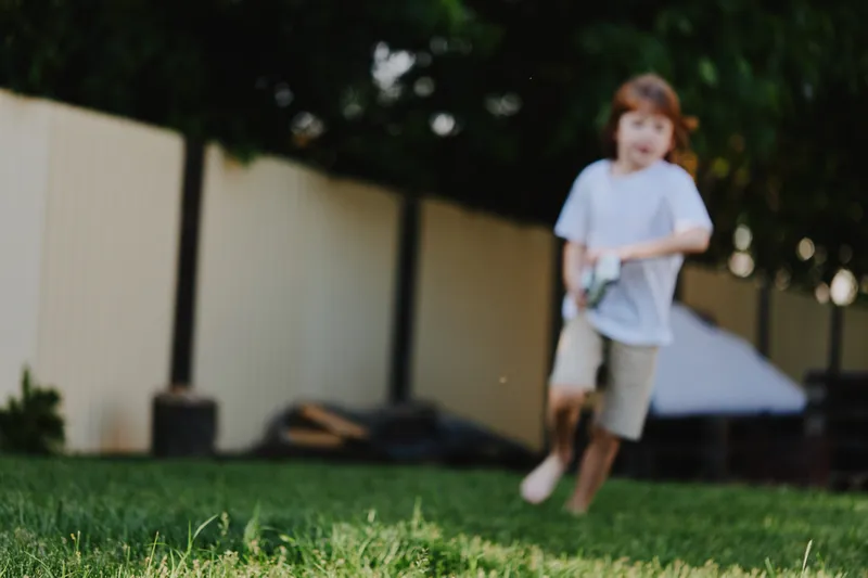 A joyful boy running through a green backyard, wearing casual clothes, expressing excitement and playfulness. Perfect for family and childhood themes.