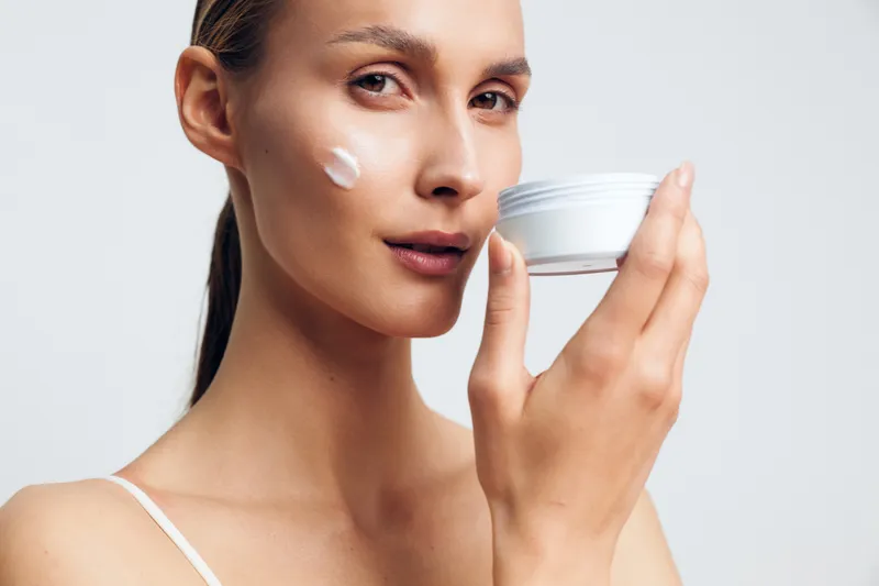 Smiling woman with a dab of cream on her cheek applying moisturizer. Close-up on a clean light background. Skincare routine, selfcare, and wellness themes. Soft studio lighting creates a gentle,