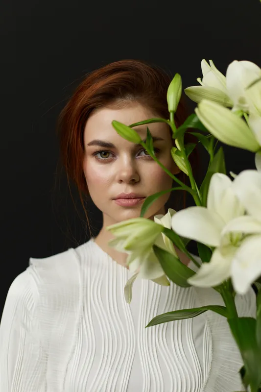 Portrait of a young woman with reddish brown hair, wearing a white blouse, gently holding elegant white lilies against a dark background, conveying beauty and freshness
