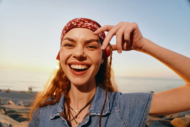 lifestyle woman wearing denim jacket and red bandana smiling outdoors with film color tones in modern boho-western street style near sunset beach