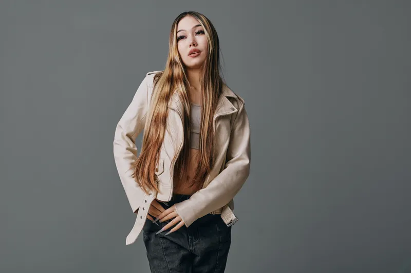 Young woman with long braided hair wearing beige jacket and black pants posing confidently against gray background in studio for fashion portrait.