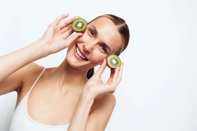 Smiling woman holding two halves of fresh kiwi near face, bright studio lighting, white background, healthy lifestyle, natural beauty, fresh fruit, joyful expression