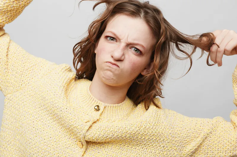 Frustrated young woman in yellow sweater pulls her hair with a tense expression on a light background. Concept of stress, anxiety, and emotional struggle in everyday life.