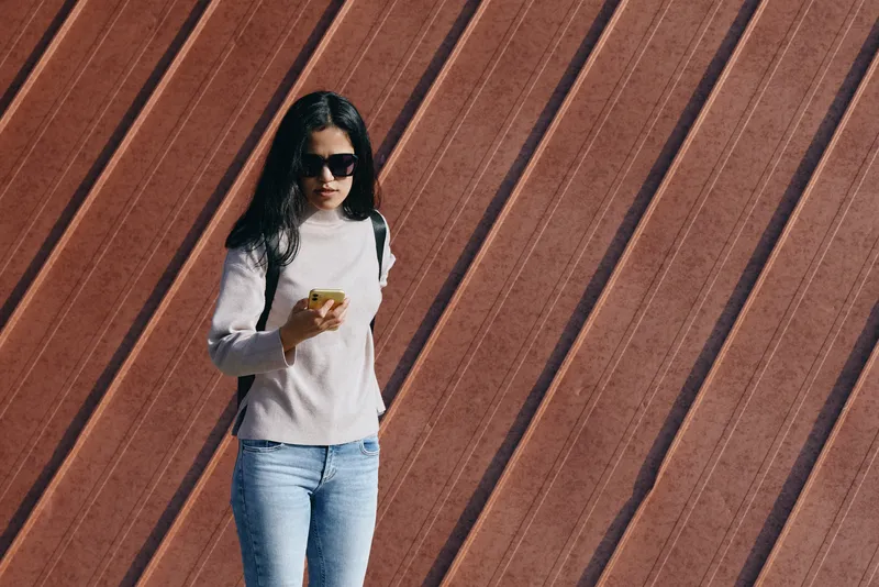 Woman wearing sunglasses and casual outfit using smartphone against diagonally angled wooden wall, urban style, modern day fashion, candid street moment outdoors, relaxed pose
