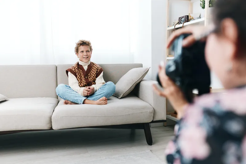 Authentic play scene featuring a young boy sitting cross legged on a sofa, dressed in playful school style, as a photographer captures candid moments at home.