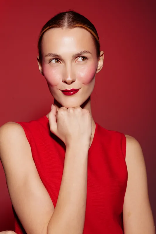 Portrait of a woman with red lipstick and blush wearing a red sleeveless top on a red background. She looks to the side with a thoughtful expression and hand near her chin.
