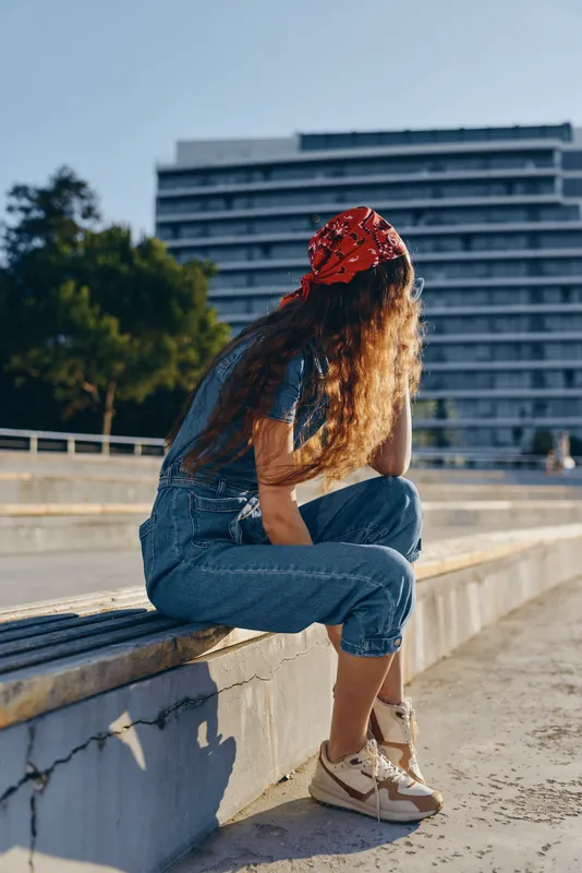 Lifestyle woman in modern boho-western denim street style sitting outdoors with natural sunlight and a red bandana, captured in warm film color tones.