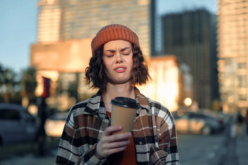 Woman holding coffee with disgusted face in urban morning, beanie and plaid jacket. Candid lifestyle portrait with golden hour glow, authenticity, mindful living and emotional storytelling.
