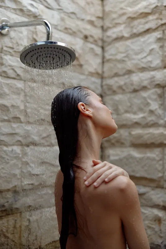 woman showering outdoors with wet hair, natural stone wall background, calm and peaceful mood, morning light bathing skin in soft glow