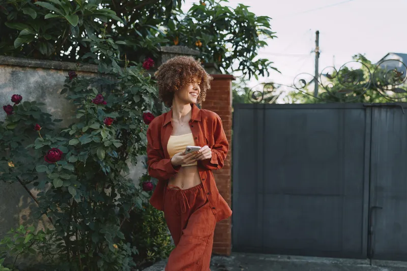 Curly haired woman in a rust suit walks along a garden path, holding a notebook, smiling. Natural daylight, blooming roses, and a casual lifestyle mood