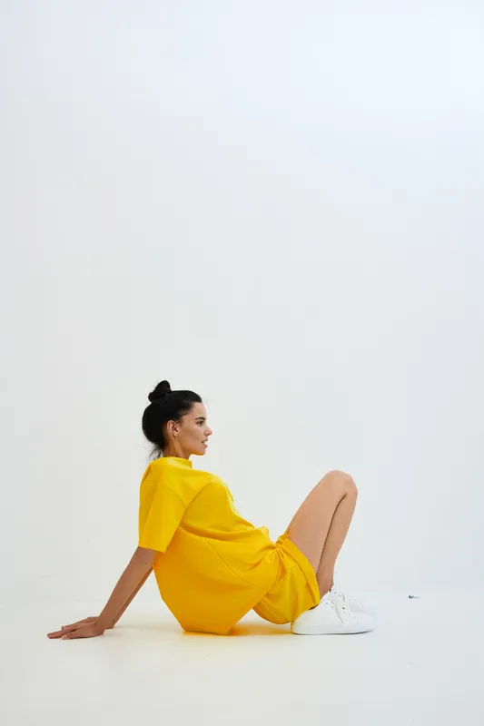 Young woman in yellow outfit sitting on white floor with relaxed pose and casual white sneakers, minimalistic background creating calm and bright atmosphere.