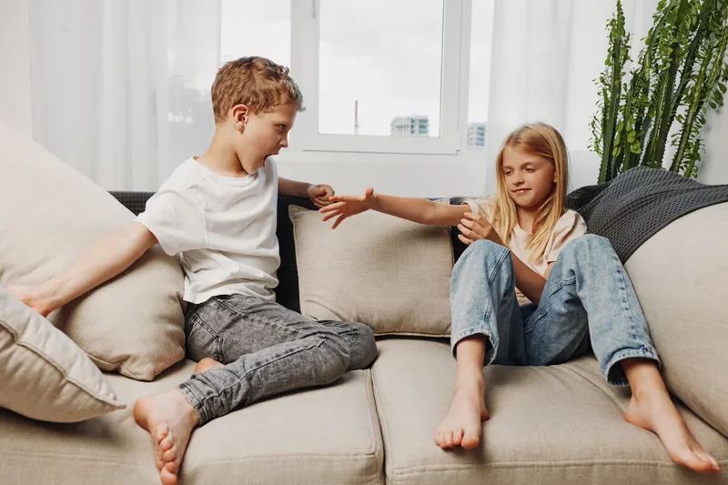 Two children sitting barefoot on a beige couch at home, interacting and playing together in a bright living room with natural light.