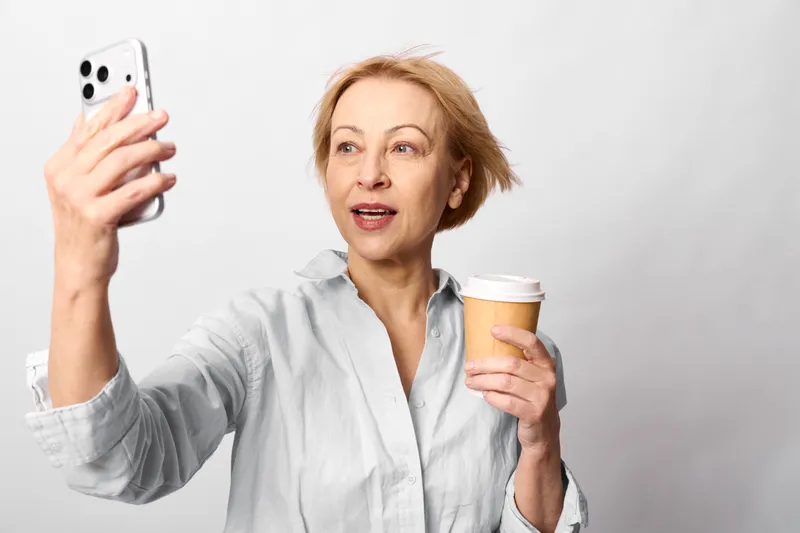 Woman takes a selfie with a smartphone, holding a coffee cup in a calm studio portrait with a white neutral background, technology and communication for social lifestyle
