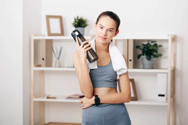 Woman in athletic wear with a towel over her shoulder holds a water bottle in a home gym, ready for a workout. Clean shelves and plants in the background create a calm fitness scene.