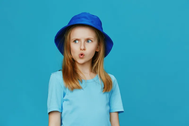 A young girl with long hair in a blue bucket hat, making an expressive face against a solid blue background, embodying playfulness and curiosity