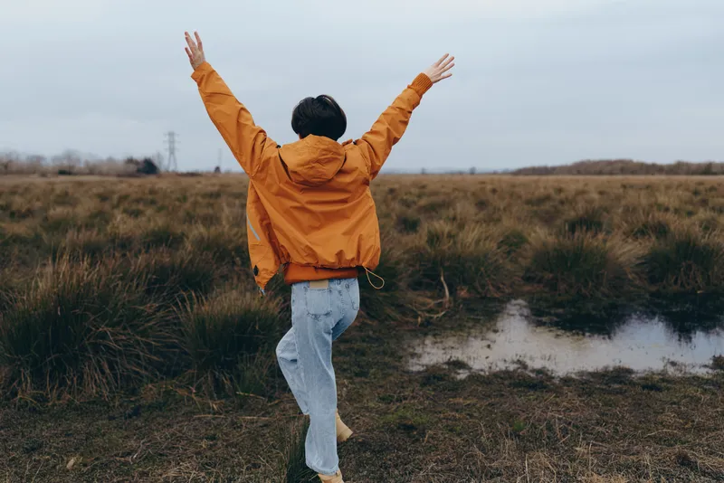 Woman lifestyle scene, outdoors in nature, arms up, wearing orange jacket and jeans, standing on a rural path near a small water puddle, cloudy sky, calm morning mood