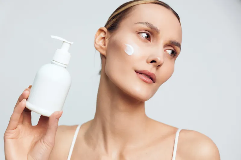 A young woman with clear skin applies white cream on her cheek holding a pump bottle. The image represents skincare, beauty routine, moisturizing, and facial care concepts.