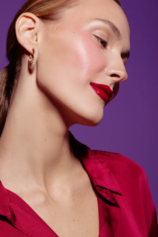 Close-up of a woman with glowing skin and red lipstick wearing gold earrings and a red blouse against a purple background. Concept of beauty, makeup, and fashion.
