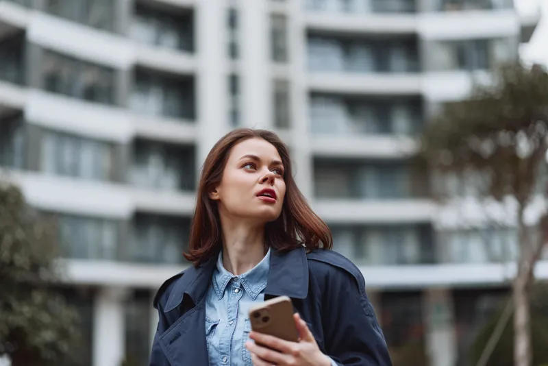 Confident woman outdoors, looking up, holding a smartphone, casual coat and denim, windy daytime, urban backdrop, natural light, soft colors, modern lifestyle moment