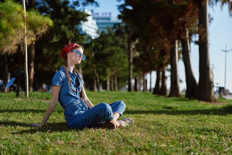 young woman sitting on grass in park wearing blue denim outfit and red headband with sunglasses enjoying sunny day outdoors relaxing near trees