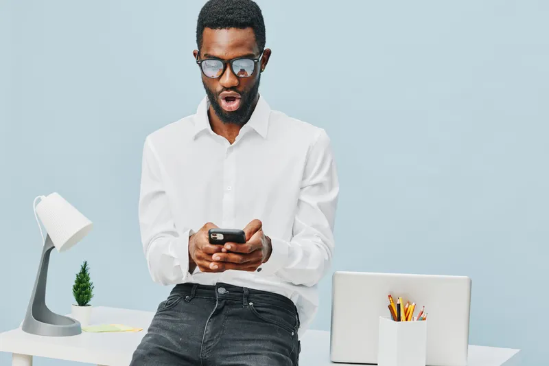 Modern man using smartphone in a bright workspace with minimal decor, wearing casual white shirt and glasses, focused on the device. Office lifestyle concept, technology, communication, casual work at