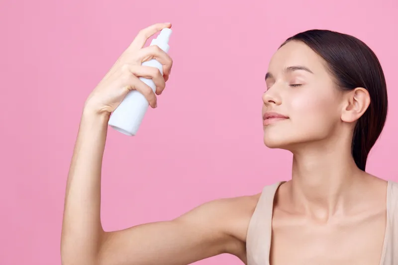 Fresh skincare routine with a young woman applying facial mist on her face against a pink backdrop The image highlights beauty, self care, and skincare products