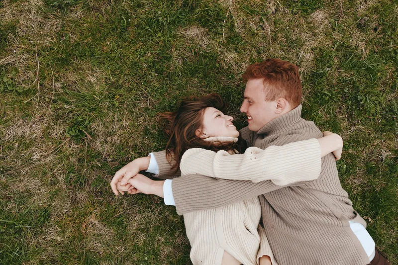 Young couple lying on grass embracing and smiling at each other in cozy sweaters enjoying a relaxed moment outdoors in natural surroundings during autumn.