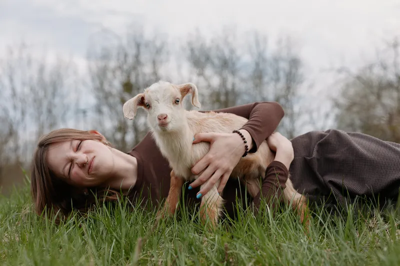 Happy woman lying on grass outdoors, cuddling a goat kid, showcasing a warm lifestyle moment, affectionate bond with animals, serene countryside vibe, relaxed smile