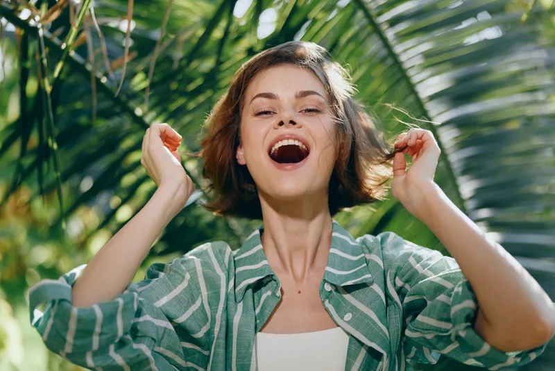 Joyful young woman outdoors, smiling broadly with arms raised, framed by tropical greenery and warm sunlight.